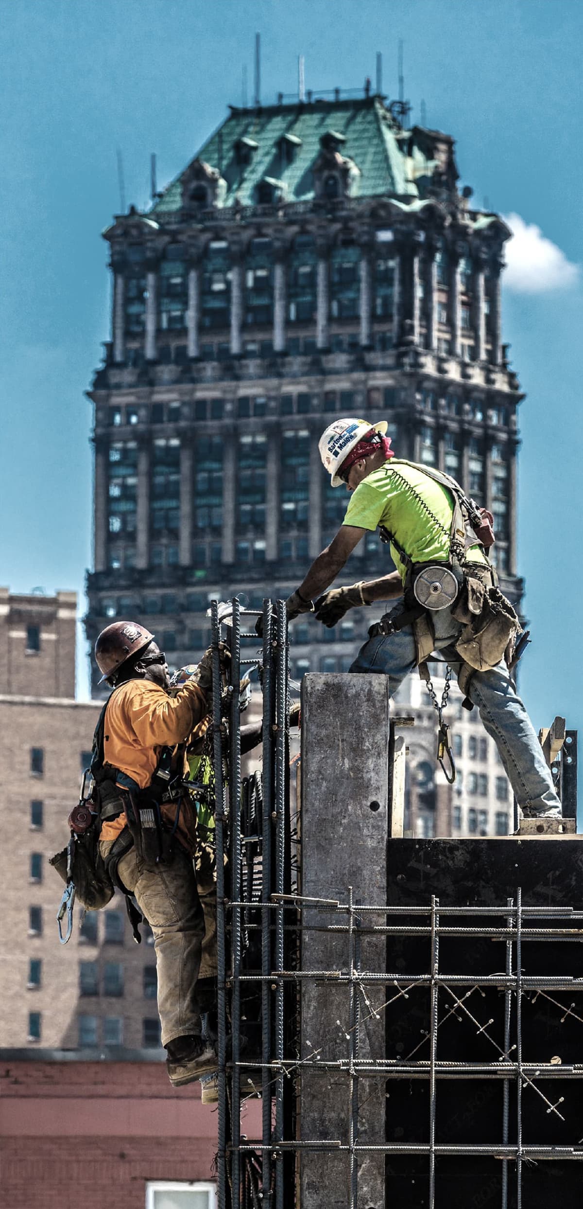 Workers tying rebar into a column form at the top of building under construction against the Book Tower building in the backgoundd. 