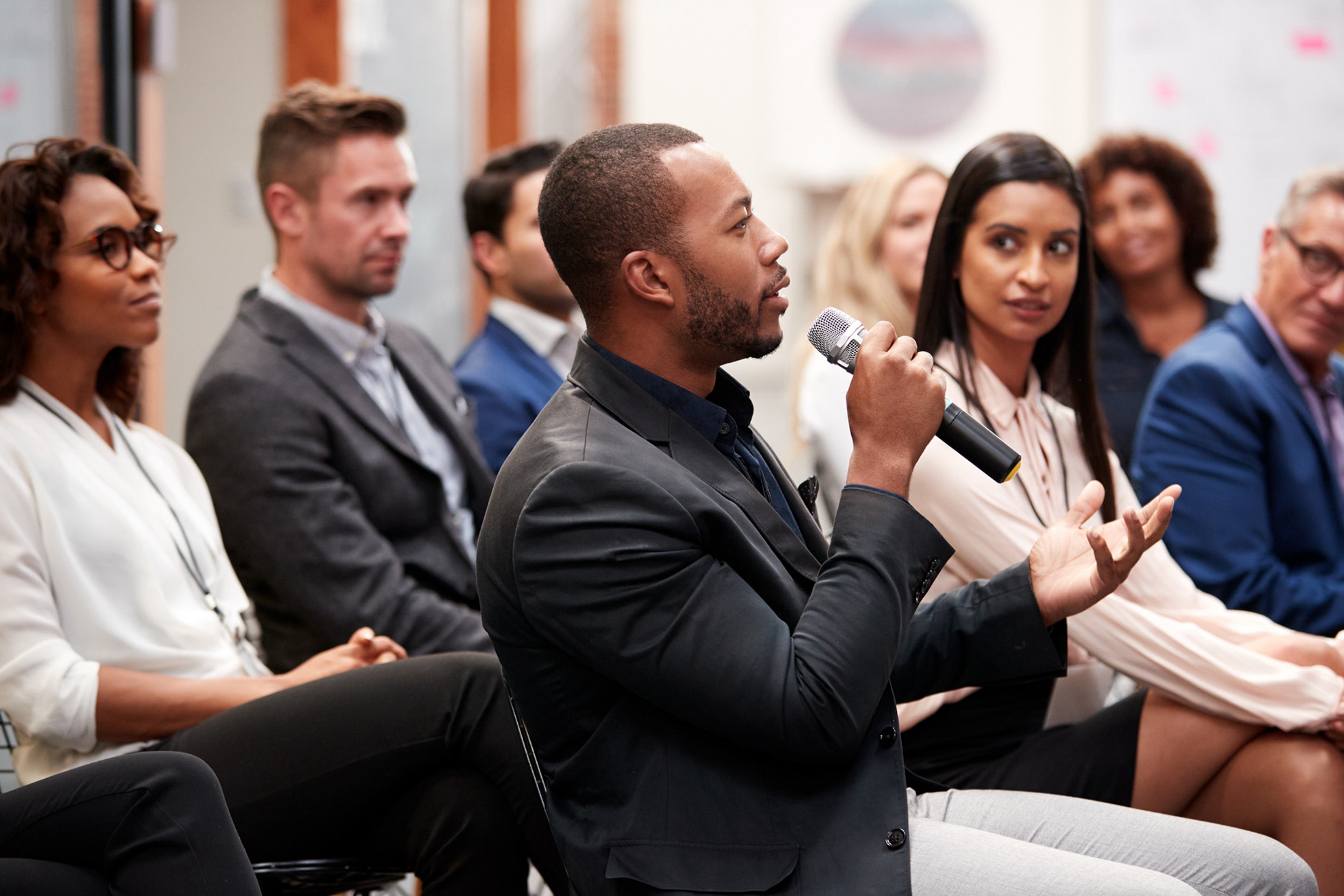 An african american man holding a mic and speaking in a group of people sitting down.